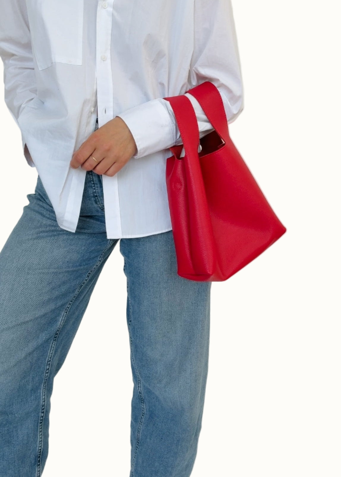 A person wearing a white shirt and blue jeans is holding a red mini tote bag with a shoulder strap.