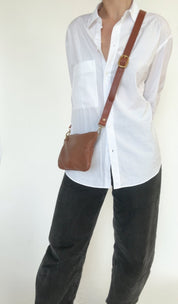 Person wearing a white shirt and black pants with a brown crossbody bag on a plain background